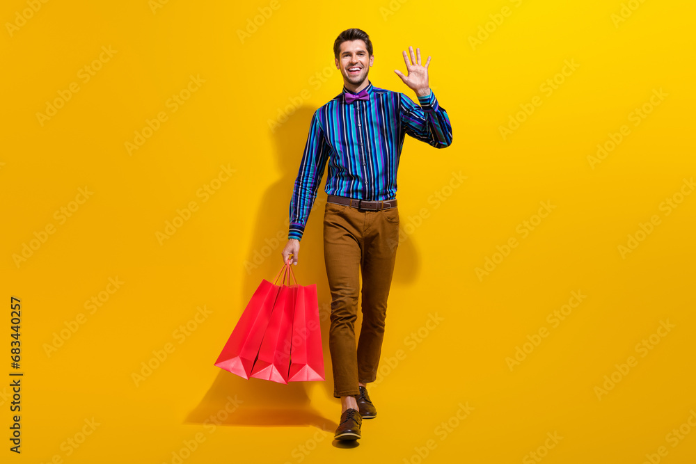 Full body photo of good mood man wear vintage bow tie stylish shirt hold shopping bags waving palm isolated on yellow color background