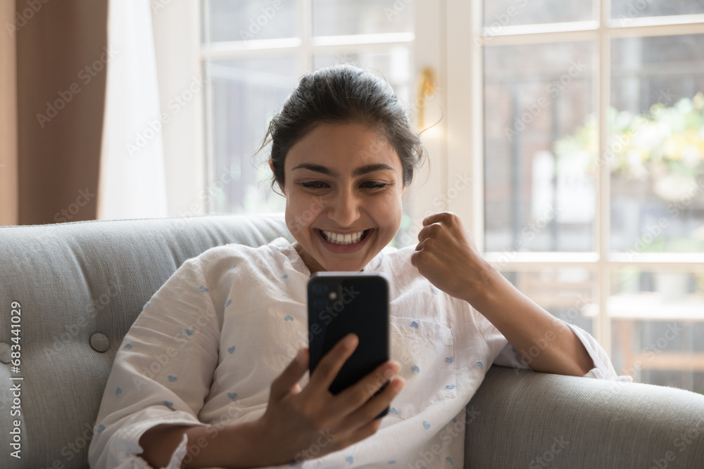 Happy Indian woman sit on sofa holds cellphone read sms, got unbelievable fantastic news feels overjoyed, rejoicing on-line lottery win, auction or bet victory, moment of success, advancement concept Happy Indian woman sit on sofa holds cellphone read sms, got unbelievable fantastic news feels overjoyed, rejoicing on-line lottery win, auction or bet victory, moment of success, advancement concept