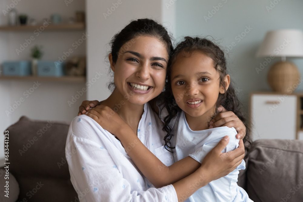 Smiling lovely little girl cuddles her young mother seated on couch staring posing for camera. Portrait of beautiful Indian happy well-being family, motherhood and custody, unconditional love concept Smiling lovely little girl cuddles her young mother seated on couch staring posing for camera. Portrait of beautiful Indian happy well-being family, motherhood and custody, unconditional love concept