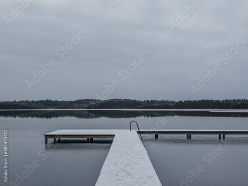A wooden footbridge covered with snow, swimming place with stairs with first ice, cloudy winter day.