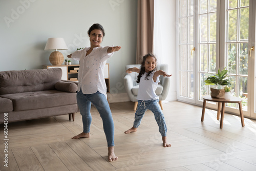 Young Indian woman and little girl perform asanas at modern home. Mom teach daughter yoga do together Warrior one exercise, standing barefoot in cozy warm living room. Healthy lifestyle, sport concept photo