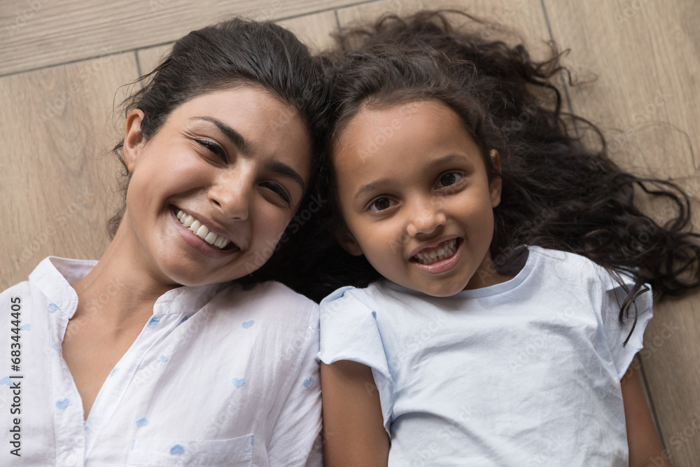 Φωτογραφία Young beautiful Indian mother and little preschooler 6s daughter lying together on warm floor smile staring at camera feel happy, close up shot