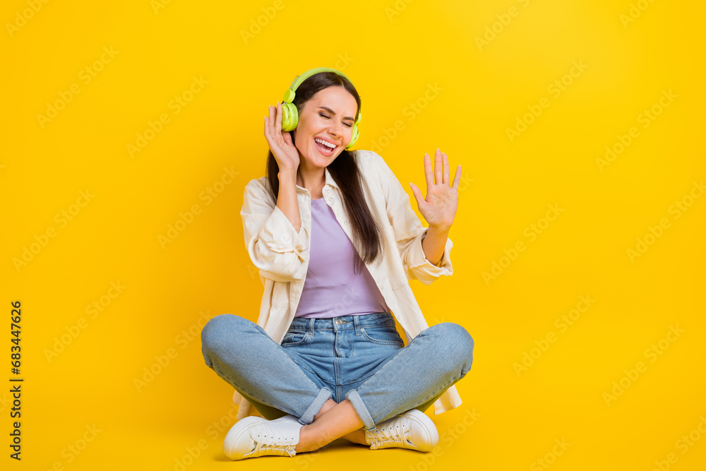 Full size portrait of pretty cheerful girl sit floor hand touch headphones dancing isolated on yellow color background Full size portrait of pretty cheerful girl sit floor hand touch headphones dancing isolated on yellow color background