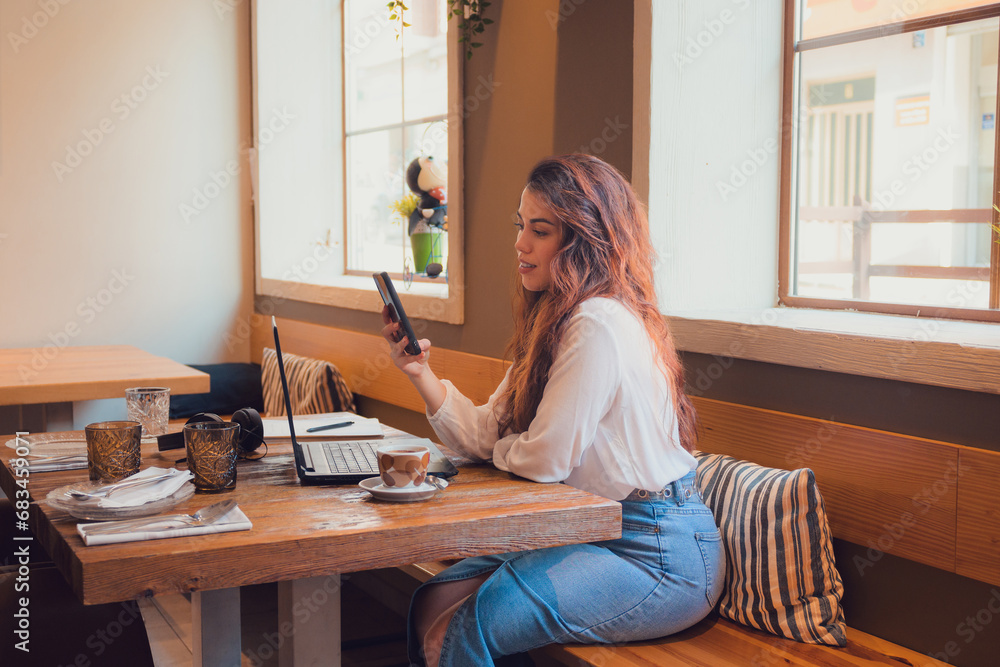 Smiling woman looking at her phone while she is working from a restaurant with a laptop Smiling woman looking at her phone while she is working from a restaurant with a laptop