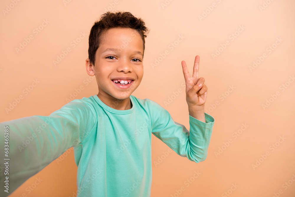 Portrait of little cheerful schoolchild toothy smile make selfie demonstrate v-sign isolated on beige color background Portrait of little cheerful schoolchild toothy smile make selfie demonstrate v-sign isolated on beige color background