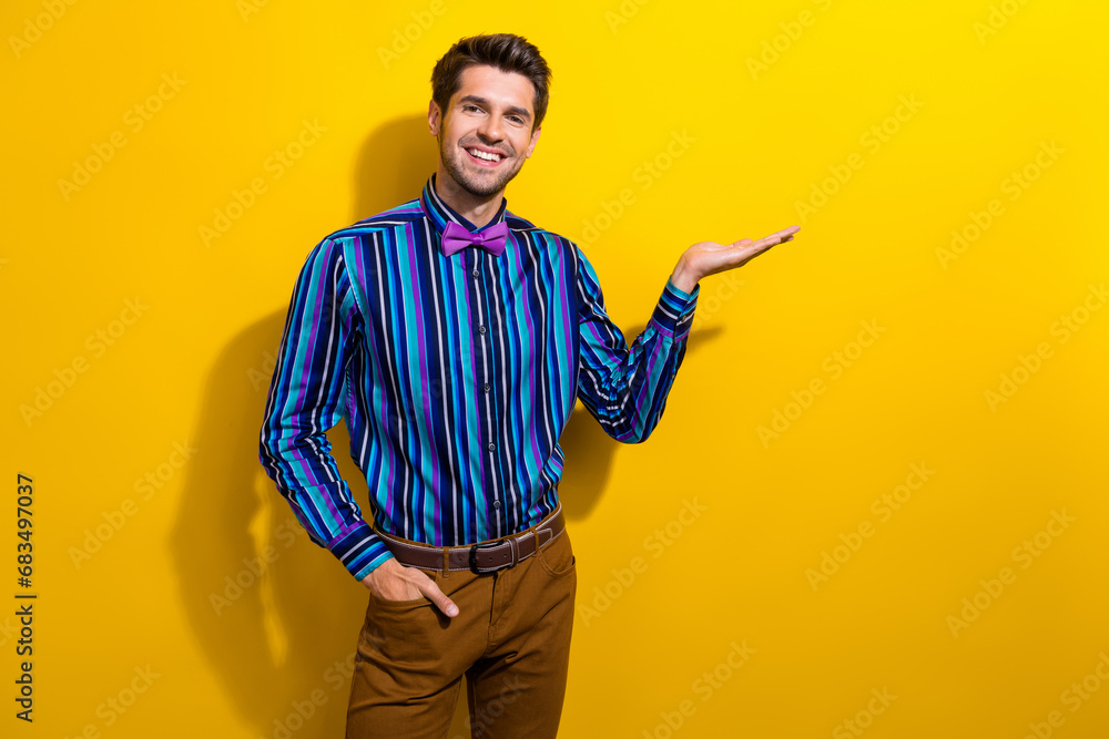 Portrait of toothy beaming guy dressed stylish shirt bow tie palm demonstrate object empty space isolated on yellow color background Portrait of toothy beaming guy dressed stylish shirt bow tie palm demonstrate object empty space isolated on yellow color background