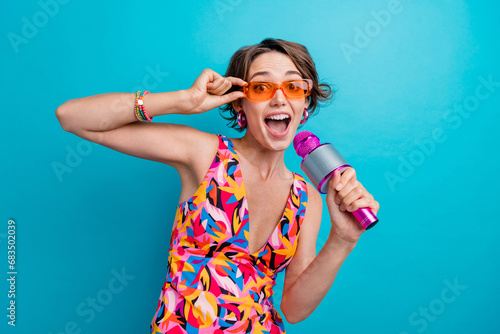 Portrait of astonished girl with short hairstyle wear colorful dress hold microphone touching sunglass isolated on blue color background