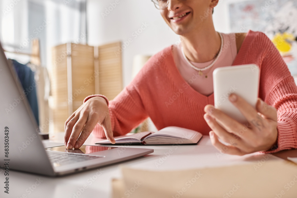 cropped androgynous freelancer in pink sweater and eyeglasses holding smartphone and using laptop cropped androgynous freelancer in pink sweater and eyeglasses holding smartphone and using laptop