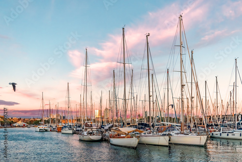Yachts moored in the Mediterranean Sea near the Port Vell promenade in Barcelona at sunset, Spain. Many boats with masts for sails against a background of pink and blue sky at dusk photo