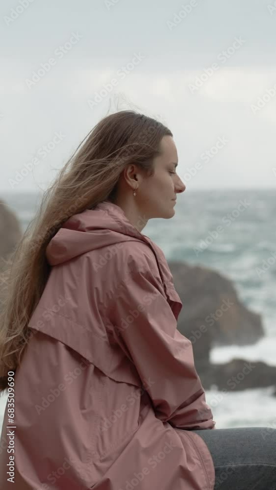 Vertical video. Solitary Young Woman in a Bad Mood Sits on Rocks by the Stormy Sea, Waves Crashing Against the Rocks, and Strong Wind Tossing Her Long Hair. Concept of Grief and Distress. Vertical video. Solitary Young Woman in a Bad Mood Sits on Rocks by the Stormy Sea, Waves Crashing Against the Rocks, and Strong Wind Tossing Her Long Hair. Concept of Grief and Distress.