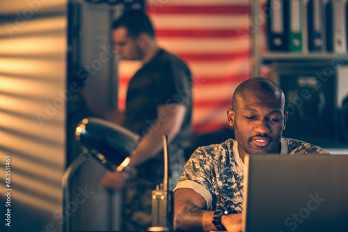 American soldier using a laptop while deployed in an army base with his roomate behind him photo