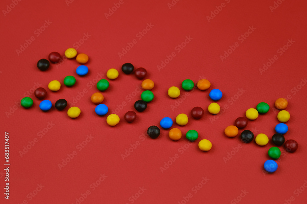 Obraz premium Happy New Year red background, inscription 2024 laid out of round chocolate colored candies. Minimalistic Christmas greeting card.