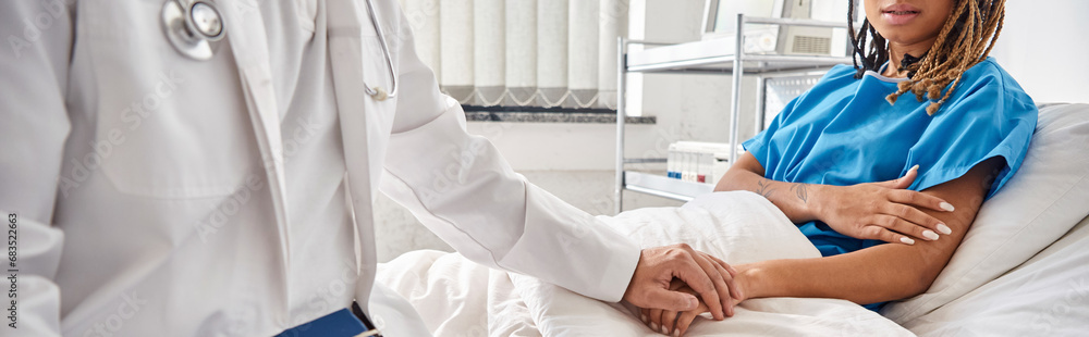 Obraz premium indian doctor holding hand of african american patient lying in hospital bed, cropped, banner