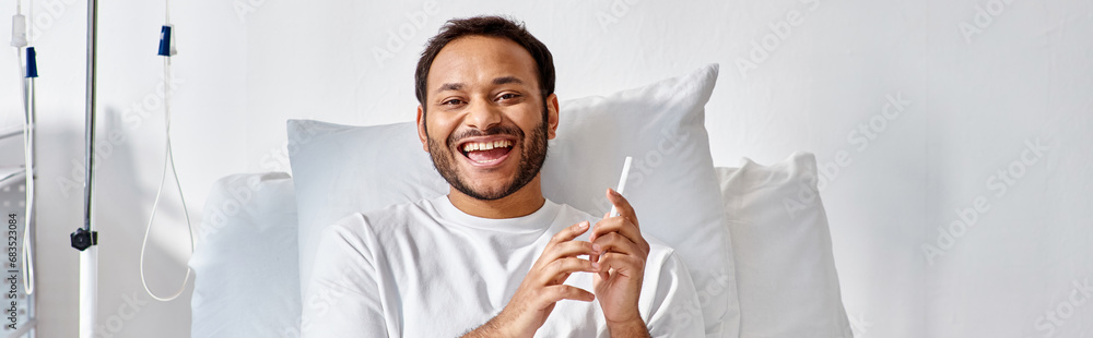 joyful indian man lying in hospital bed with phone in hands and smiling cheerfully, banner joyful indian man lying in hospital bed with phone in hands and smiling cheerfully, banner