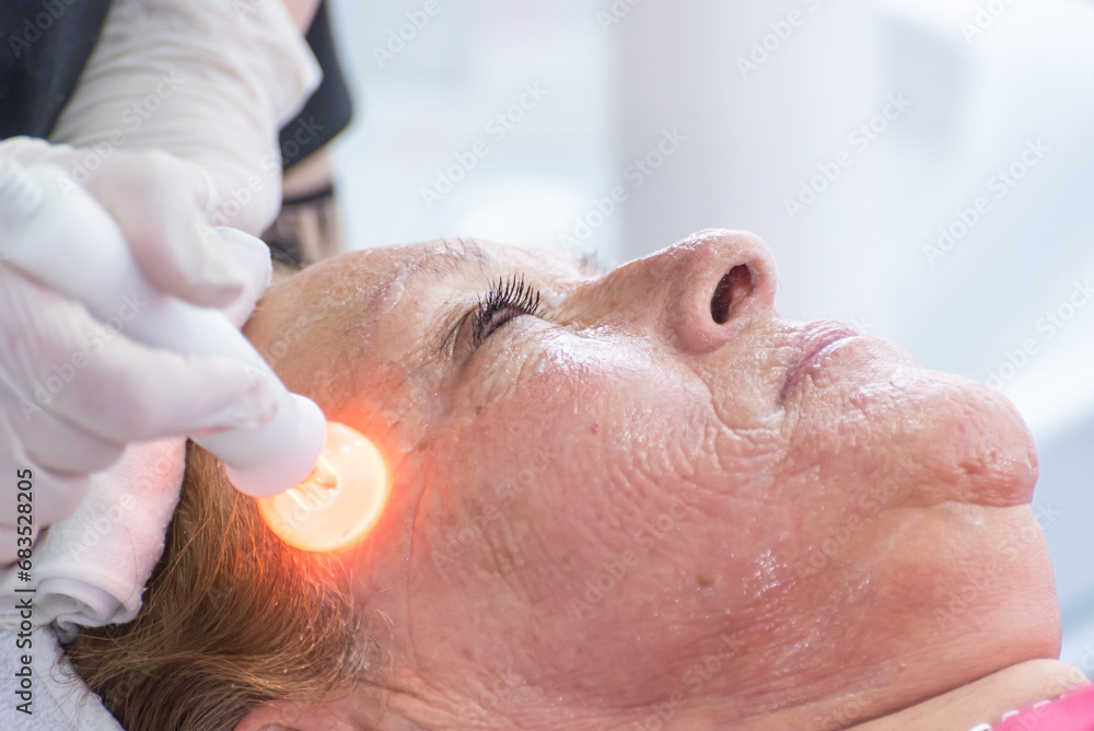Naklejka premium Beautician performing phototherapy with LED light to adult woman in SPA salon, facial phototherapy to clean pores of the skin. Anti-aging and photo rejuvenation treatments. facial treatment.