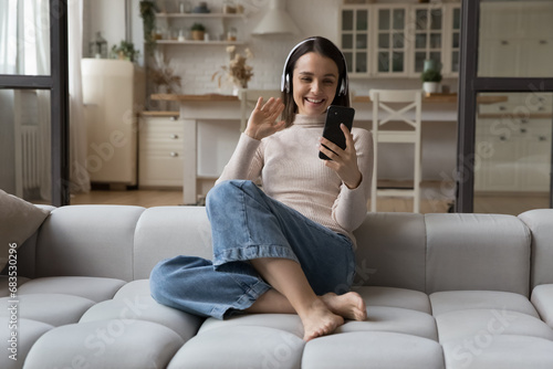 Young smiling woman in headphones greets friend or family living abroad use modern smartphone and video conference app, sits on cozy sofa at home. Virtual meeting event, remote communication concept
