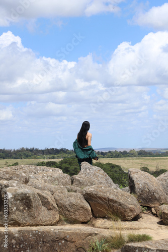 Mujer joven de pelo largo negro mirando el horizonte de espaldas a camara caminando entre las rocas de la montaña en un dia soleado