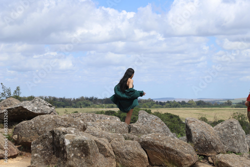Mujer joven de pelo largo negro mirando el horizonte de espaldas a camara caminando entre las rocas de la montaña en un dia soleado
