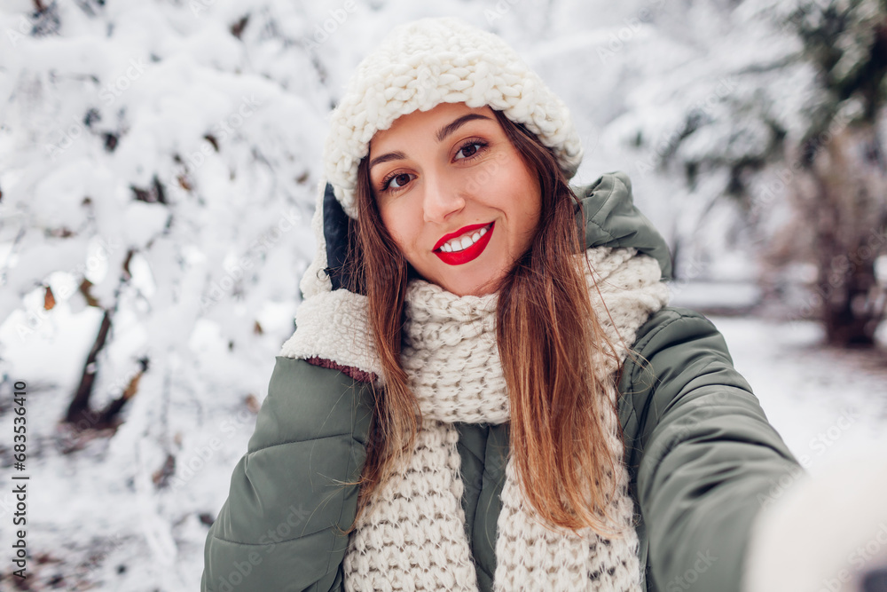 Portrait of young smiling woman taking selfie in snowy winter park wearing warm knitted hat scarf mittens. Close up Portrait of young smiling woman taking selfie in snowy winter park wearing warm knitted hat scarf mittens. Close up