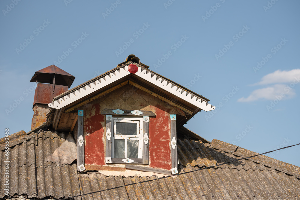 An old attic with a window and a chimney outside a dilapidated house against a blue sky.