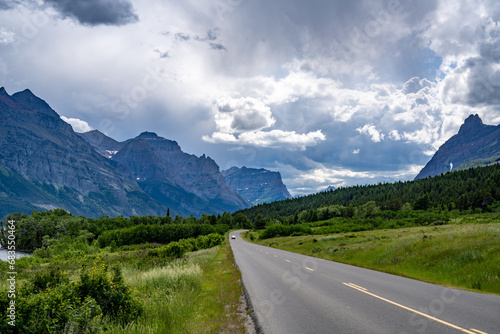 A road curving through a grassy slope, forest and rugged mountains under dramatic clouds, Going to the Sun Road, Glacier National Park, Montana