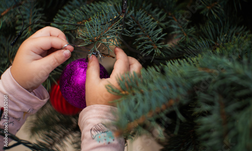 Little children's hands are trying to remove the New Year's toy from the Christmas tree. Close-up. A curious kid is trying to steal a shiny New Year's ball from the Christmas tree.