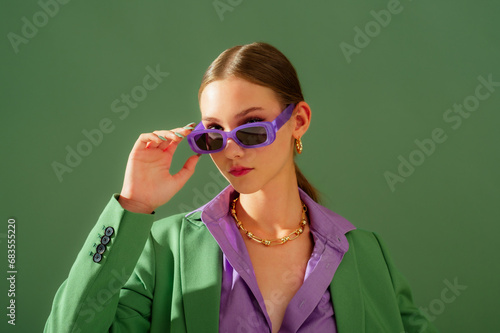 Fashionable beautiful confident woman wearing trendy purple color rectangular sunglasses, suit blazer, office shirt, chain necklace, posing on green background. Copy, empty space for text