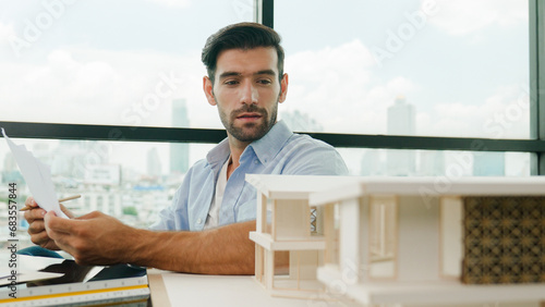 Portrait of smart skilled engineer checking house model while holding blueprint. Professional businessman choosing project while looking at architect model and thinking designing,choosing. Disputation
