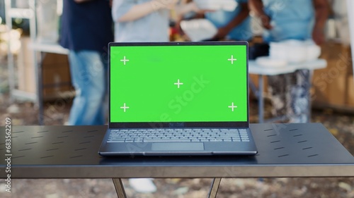 Close-up of electronic laptop with isolated green screen sits on a table while charity workers assist the less privileged. Detailed view of minicomputer with a blank copyspace mockup template.