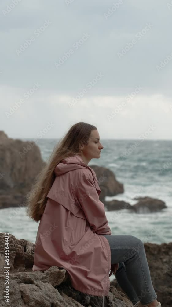 Vertical video. Young Lonely Woman Sits on Sharp Rocks in a Pink Raincoat, Gazing at the Sea with Waves and Rocks Behind Her. Vertical video. Young Lonely Woman Sits on Sharp Rocks in a Pink Raincoat, Gazing at the Sea with Waves and Rocks Behind Her.