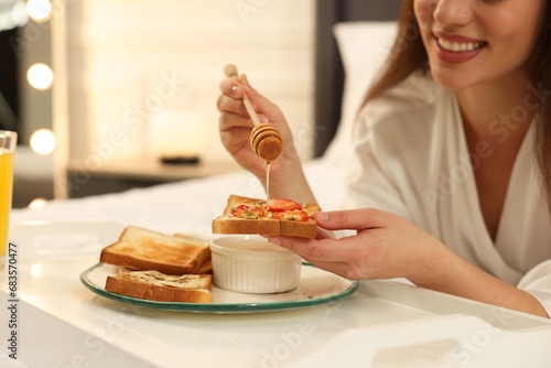 Happy young woman pouring honey onto toast near white tray on bed at home, closeup photo