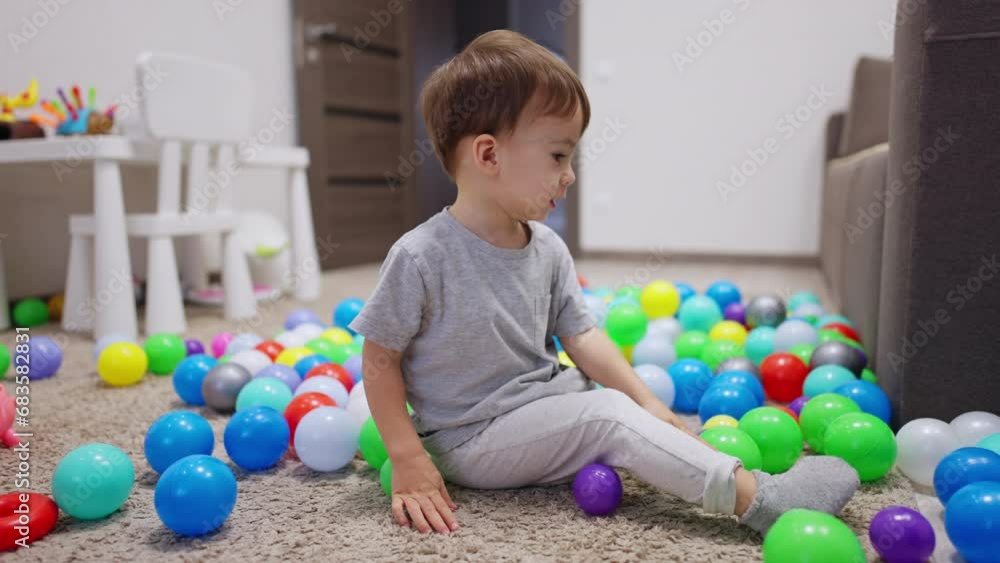 Two year old toddler sits on the floor among the colorful balls. Kid takes green ball and throws it ahead. Two year old toddler sits on the floor among the colorful balls. Kid takes green ball and throws it ahead.