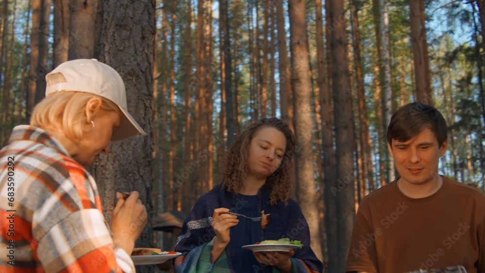 Fototapeta premium Friends eat barbecue from grill in woods. Stock footage. Friends relax in nature at barbecue and eat. Friends eat cooked meat on grill on sunny summer day