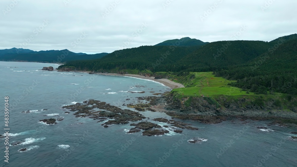 Top view of stone reefs off coast with green forests and rocks. Clip.Beautiful waves at reef shore with green forests of mountains. Northern landscape of green mountains and rocks on shore on cloudy