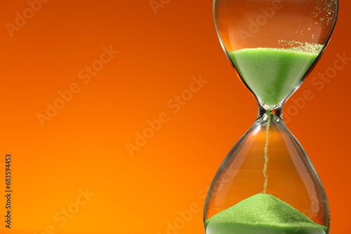 Hourglass with light green flowing sand against orange background, closeup. Space for text