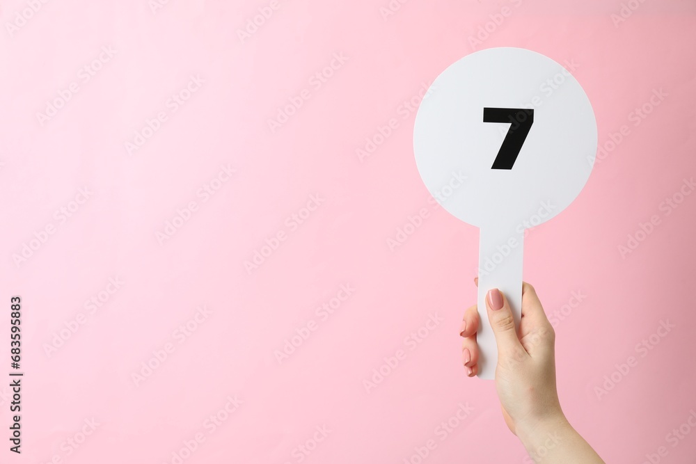 Woman holding auction paddle with number 7 on pink background, closeup. Space for text Woman holding auction paddle with number 7 on pink background, closeup. Space for text