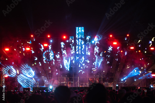 The night is alive with music and excitement at a concert festival main event. A cheering unrecognizable crowd gathers in front of brightly lit stage and the lens flare adds to the lively atmosphere.
