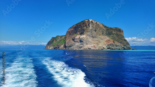 Looking at Guishan Island from a departing yacht. There are blue cloudy sky in background. Waves splashing in the sea as the boat passes by, and creating bubble foaming on water surface.