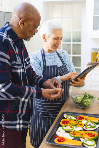Diverse senior couple preparing healthy meal with vegetables using tablet in kitchen