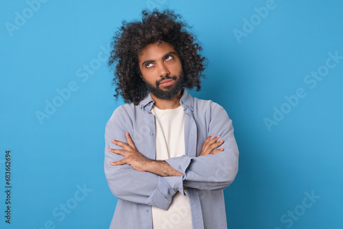 Young doubtful funny Arabian man rolls eyes and looks up with offense crossing arms in front of chest after insult or inappropriate remark from boss posing on isolated blue background.