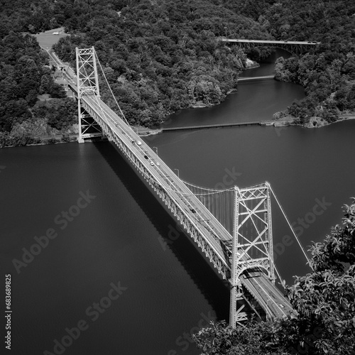 Transportation infrastructure and nature. A high-angle black-and-white view showing the Bear Mountain suspension bridge rising out of the Hudson River and a verdant woodland. Anthony's Nose, New York