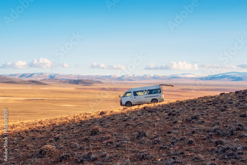 An old white van with an open rear trunk, parked on a steep mountain slope against the background of the autumn steppe, an extreme journey through the vast expanses.