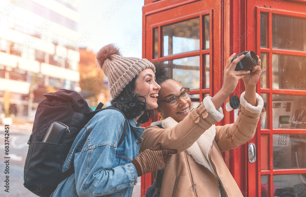Fototapeta premium two friends, girlfriend and women using a mobile phone, camera and taking a selfie against a red phonebox in a city in England. Travel Lifestyle concept