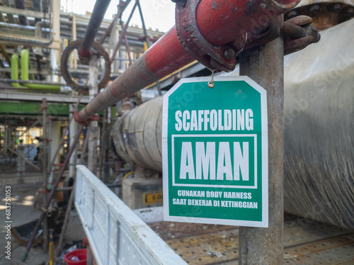 Hanged green sign label tag with writing in scaffolding aman, gunakan body harness saat bekerja di ketinggian in English safe scaffolding, use body harness when working at heigh photo