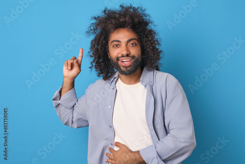 Young cheerful Arabian man student experiences stomach pain from laughing and points finger up inviting him to attend stand-up show with popular comedians stands posing in blue studio.