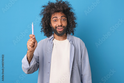Young inspired attractive Arabian man with black hair raises pen up and opens mouth wanting to tell new life hack or way to save money dressed in casual style stands on blue studio background.
