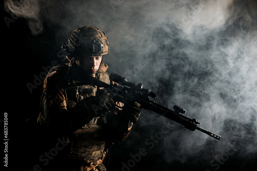 An army soldier in a military camouflage uniform, a helmet and a mask, holds a rifle and aims with a red dot sight, standing in an attack position, in the smoke on a black background. photo