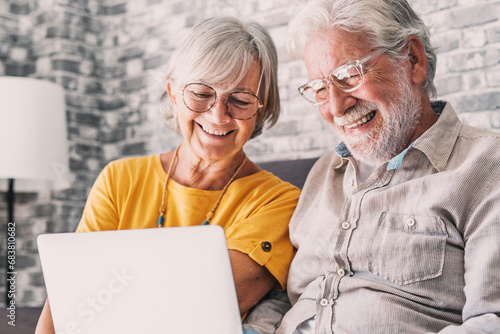 Pretty elderly 70s grey-haired couple resting on couch in living room hold on lap laptop watching movie smiling enjoy free time, older generation and modern wireless technology advanced users concept.
