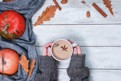 Cropped shot of young woman hands warming hot coffee mug, cup of latte, spicy cortado, bright orange pumpkins on cozy woolen scarf, dry leaves, spices on white wooden table. Autumn background