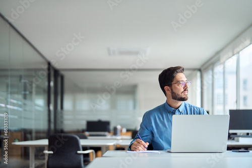 Busy young latin businessman working in office using laptop looking away. Serious professional business man manager thinking on project strategy plan ideas sitting at workplace. Copy space.
